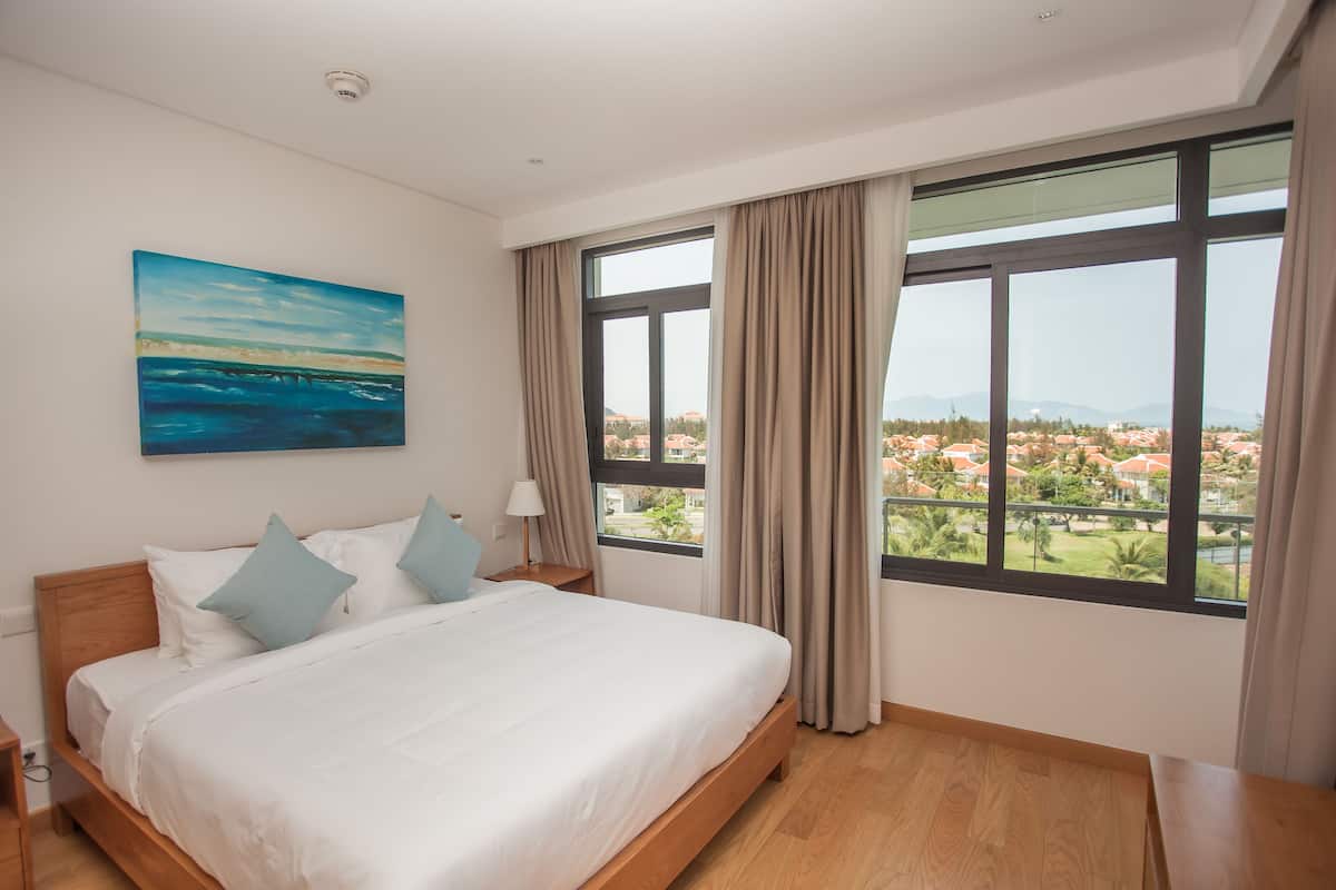 Habitación de hotel moderna con una cama grande, un cuadro de playa y vistas a un paisaje urbano a través de amplios ventanales.