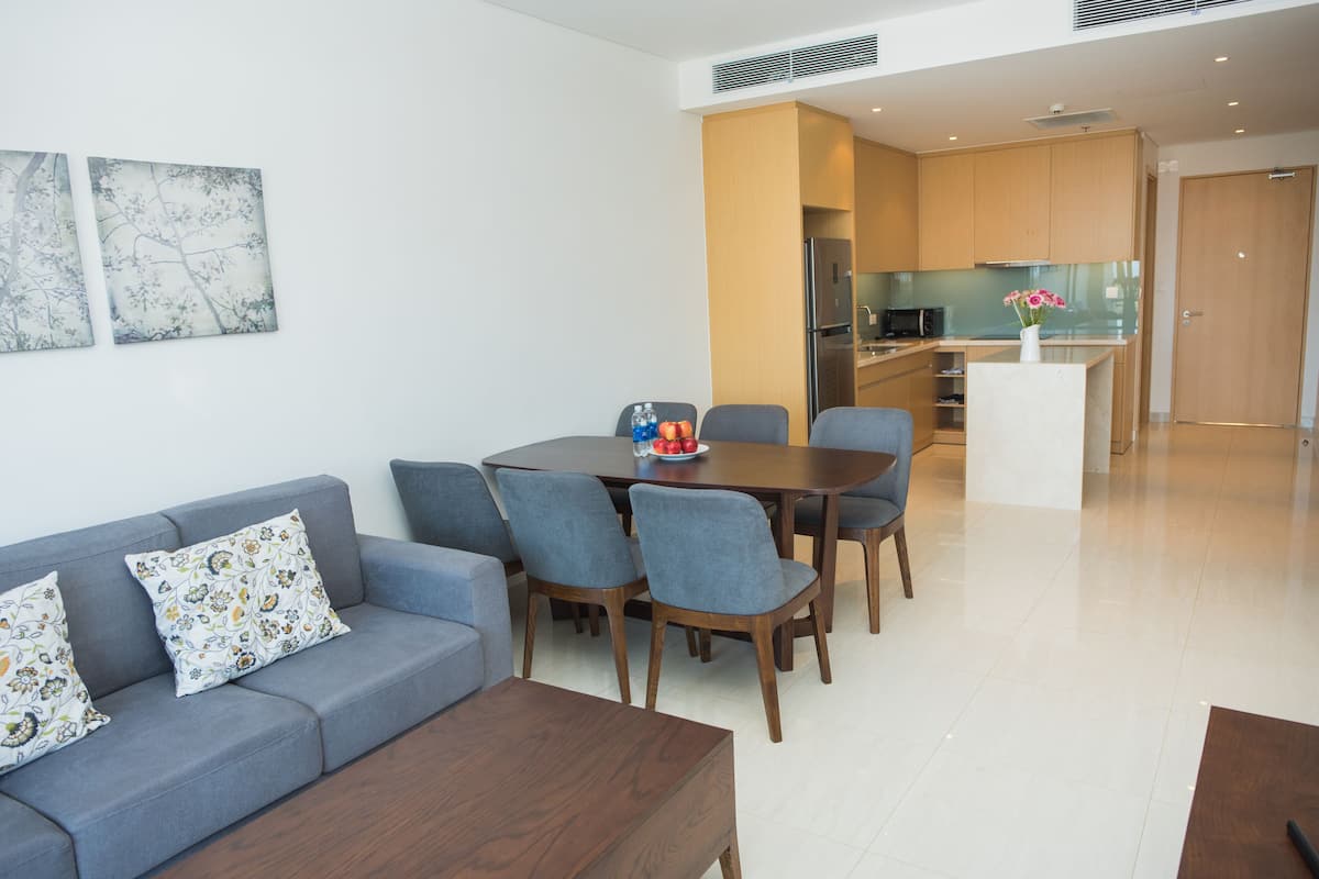 Una sala moderna con un sofá gris, una mesa de centro de madera y un comedor con una mesa y sillas de madera. Se ve una cocina al fondo.