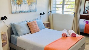 A neatly made bed with a grey frame, blue and orange pillows, and an orange blanket. There is a ceiling fan above the bed, a painting of trees on the wall, and a window with curtains. - Beautiful Caribbean Ocean View Gem ! Newly remodeled apartment.  (Luquillo)