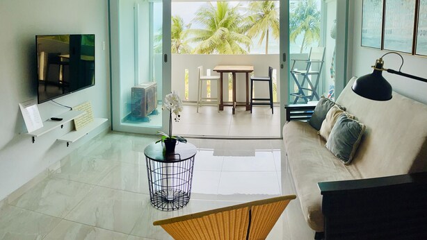 A modern living room with a flat-screen TV, a ceiling fan, a sofa, a coffee table, and a view of palm trees through the glass doors. - Beautiful Caribbean Ocean View Gem ! Newly remodeled apartment.  (Luquillo)