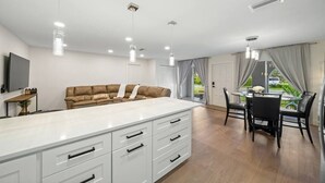 A modern kitchen with white cabinetry, a central island with drawers, pendant lighting, a brown sofa with 'HOME' on it, and a dining area with a glass table and chairs.