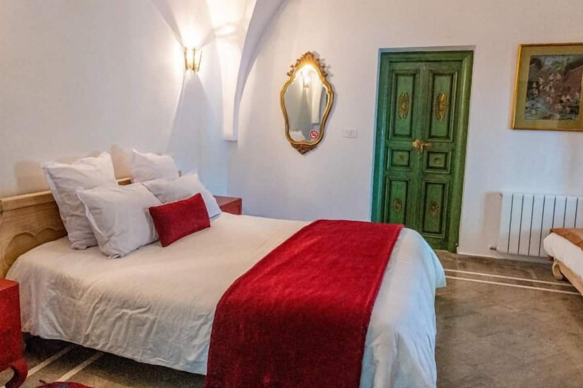 A bedroom with a bed, a nightstand, a red lamp, a green door, and a wall-mounted mirror.