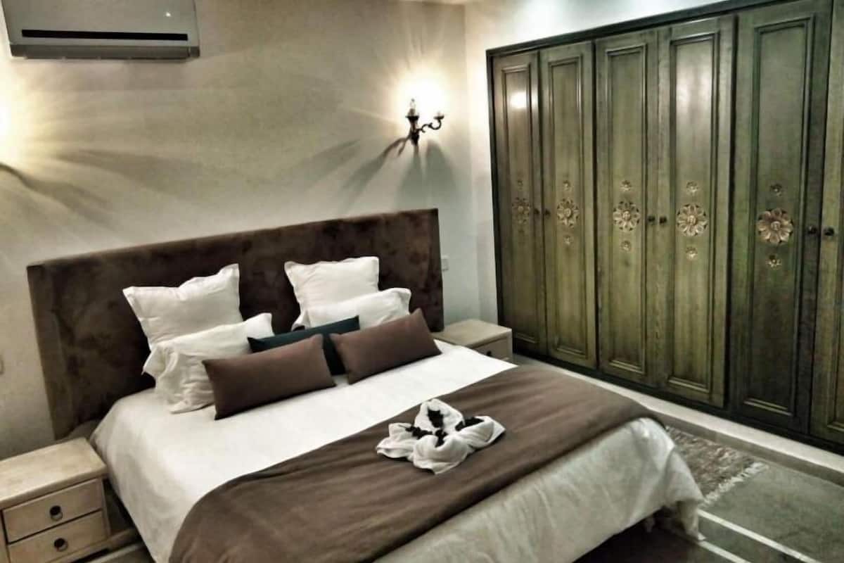 A bedroom with a large bed, a nightstand, a wardrobe, and an air conditioning unit.