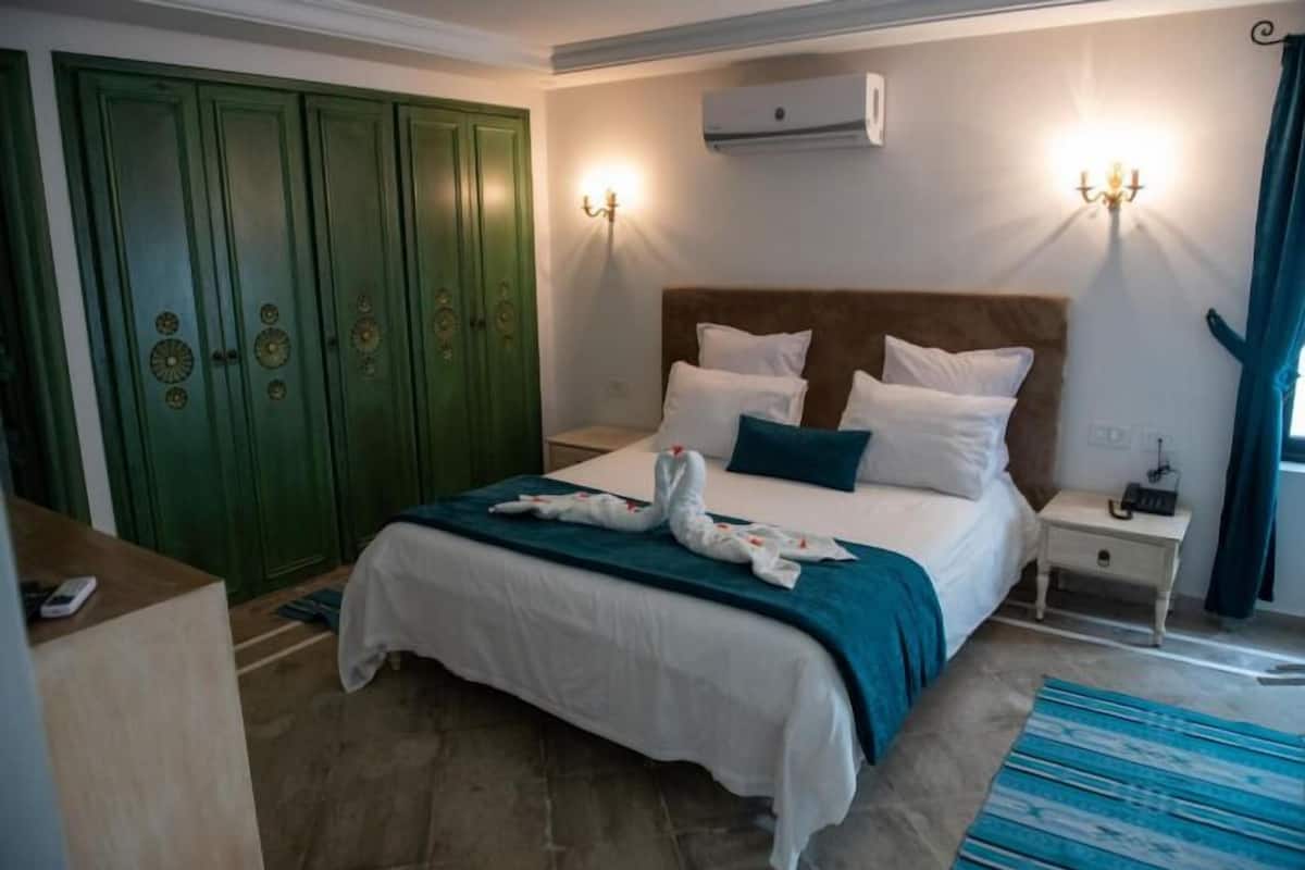 A bedroom with a bed, green wardrobe, bedside lamps, and a blue striped rug.