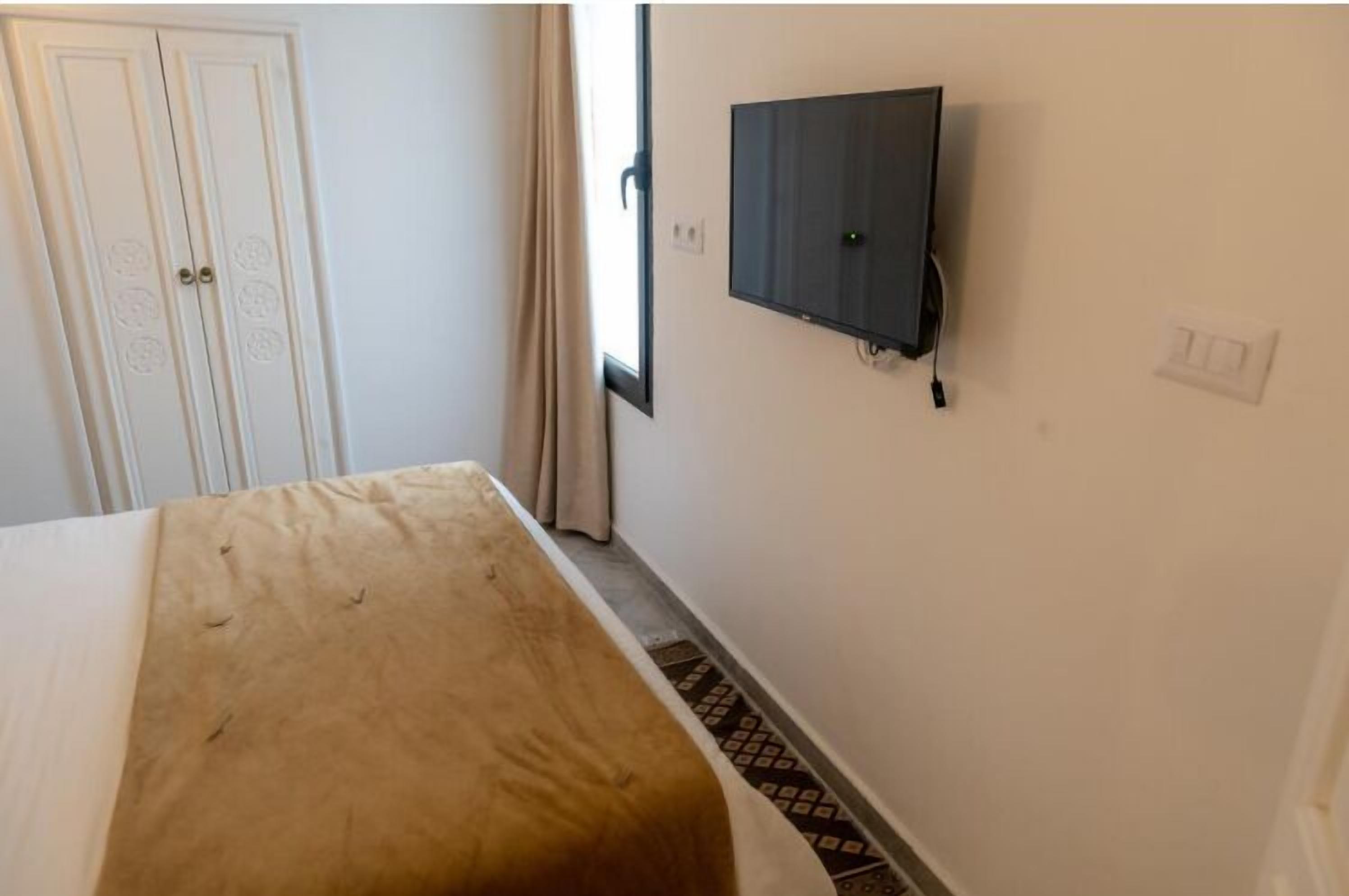 Una habitación con una cama, televisión montada en la pared, una puerta y un ventanal con cortinas.