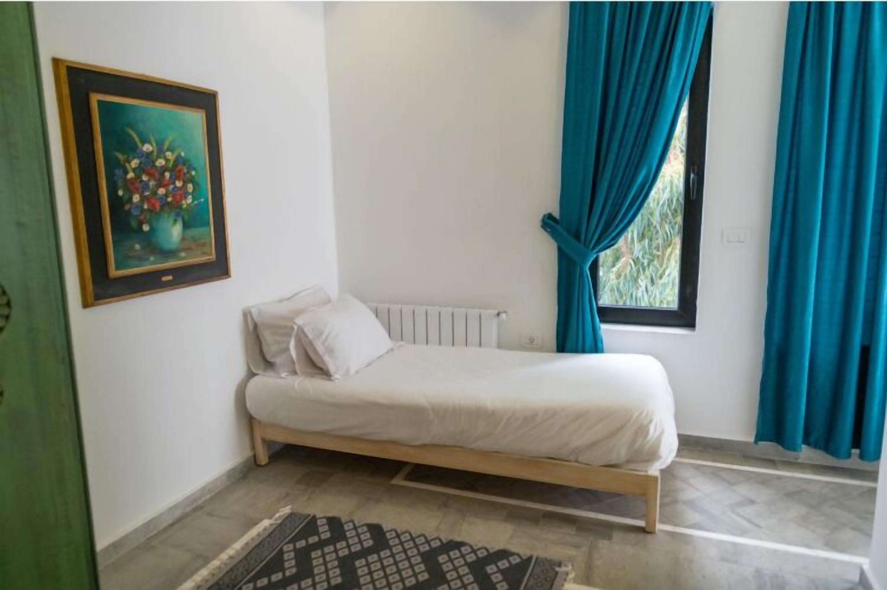 A single-bed room with a white mattress, a framed floral painting on the wall, a radiator, and teal curtains.
