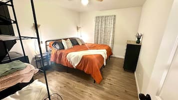 A bedroom with a bunk bed, a single bed with an orange bedspread, a ceiling fan, a window with curtains, and a small table with a vase.