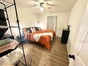 A bedroom with a bunk bed, a single bed with an orange bedspread, a ceiling fan, a window with curtains, and a small table with a vase.