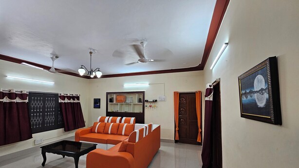 A room with a sofa, a coffee table, a ceiling fan, a framed picture, and a door with curtains. - Family Home in Prime Location close to City Center (Sathamangalam)