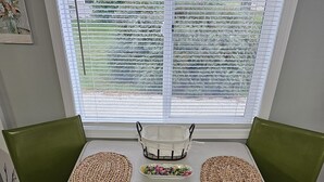Una mesa blanca con dos individuales tejidos, una cesta blanca y dos sillas verdes en una habitación con ventana a un jardín.