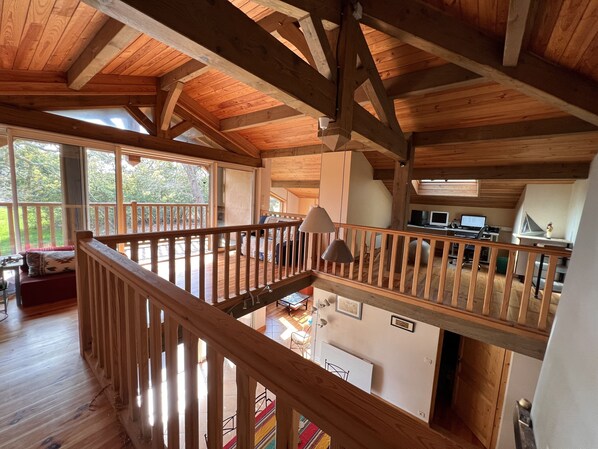 Amplia habitación estilo loft con techos altos y vigas de madera a la vista. Incluye un balcón con barandilla de madera, una zona de comedor con mesa y sillas, y una cocina con encimera y electrodomésticos.