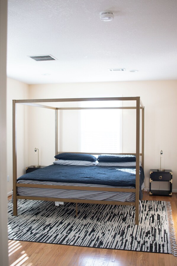 A four-poster bed with a dark blue bedspread and pillows, a black and white patterned rug, and a wooden floor. - Gorgeous Renovated Historic Home. (Houston)
