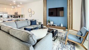 A modern living room with a grey sectional sofa, a coffee table, and a blue armchair. There is a kitchen in the background with white cabinets and a dining area. - Championsgate-Free Water park/18 mins DisneyWorld (Davenport)