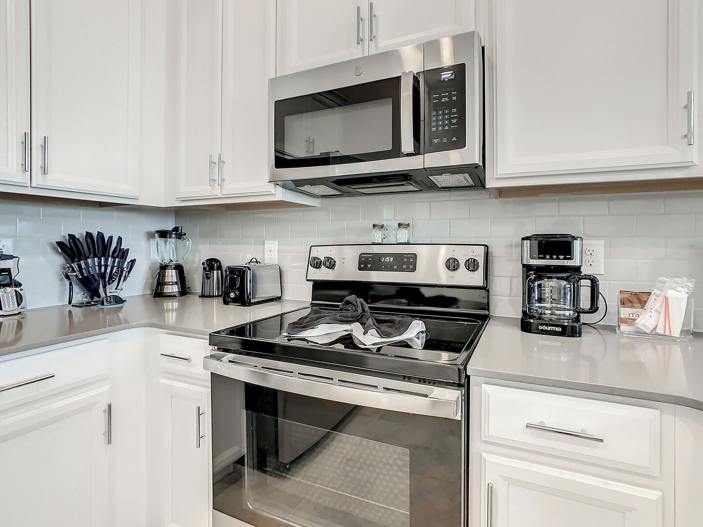Una cocina moderna con armarios blancos, electrodomésticos de acero inoxidable, incluyendo un microondas, tostadora y cafetera, y una estufa a gas.