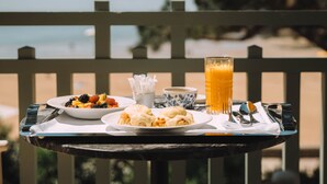 Ein Tisch mit Frühstücksangeboten, darunter Pfannkuchen, Obst und Orangensaft, befindet sich auf einem Balkon mit Blick auf den Strand.