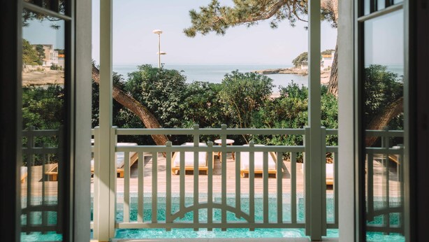 Ein bogenförmiger Balkon mit Pool, mit Blick auf eine Küstenlandschaft mit Bäumen und blauem Himmel.