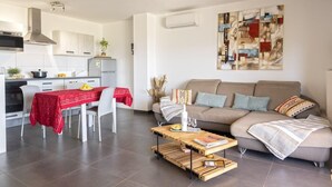 A modern kitchen with white cabinets, a dining table with a red tablecloth, and a living area with a beige sofa and abstract art on the wall. - Casa Ghju (Algajola)