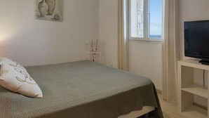 A bedroom with a bed, a television on a stand, a window with curtains, and a painting on the wall. - Casa Lucia (Algajola)