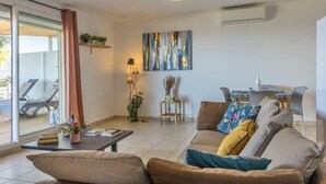 A modern living room with a sofa, a coffee table, and a dining area with a table and chairs. There is a sliding glass door leading to an outdoor area with a sun lounger. - Casa Lucia (Algajola)