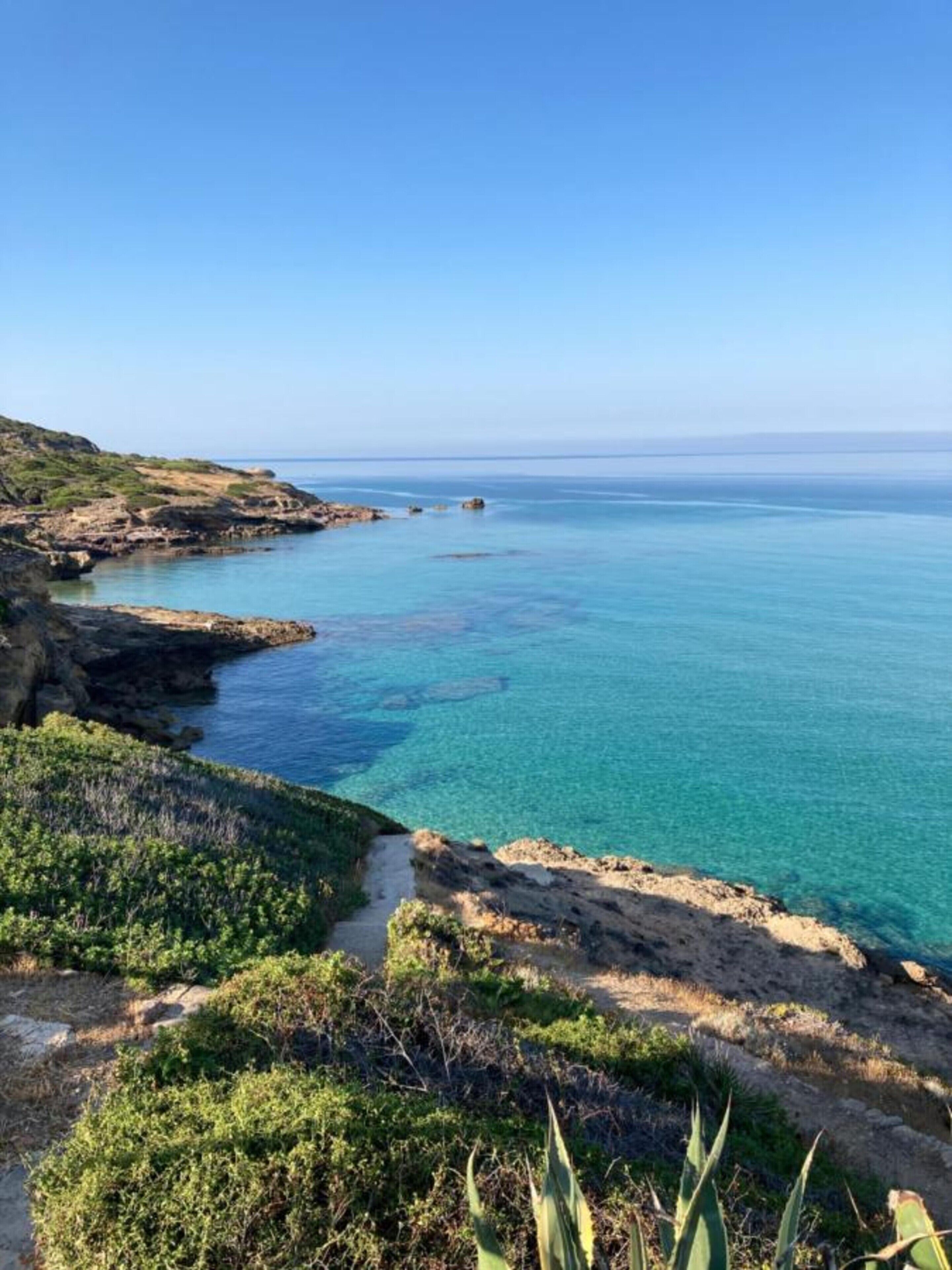 A coastal landscape with clear blue sea, rocky terrain, and sparse vegetation.