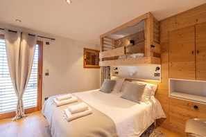 A bedroom with a bunk bed, a wooden wardrobe, a window with curtains, and a bedside table.