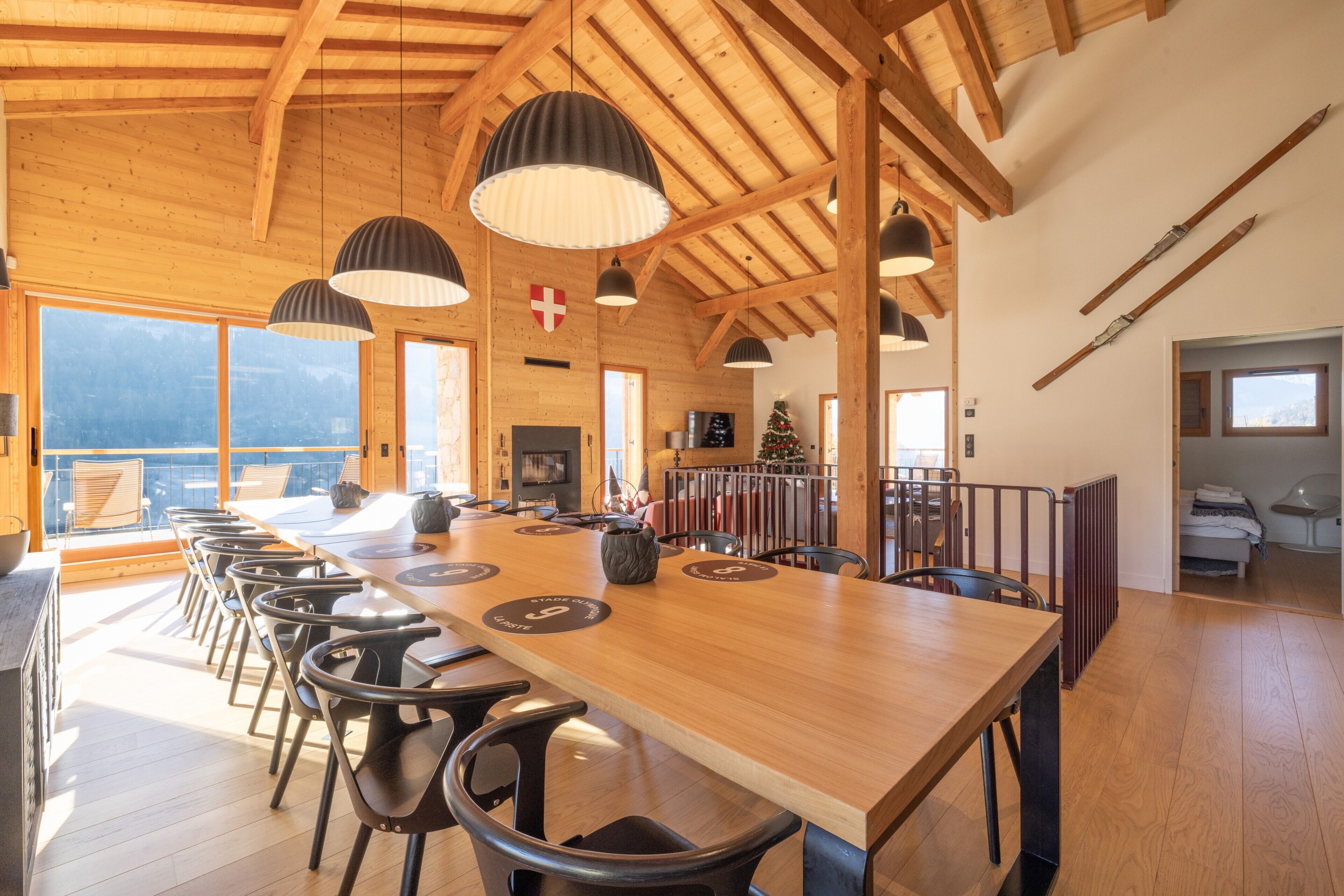 Een houten interieur met een lange tafel, stoelen, hanglampen en een uitzicht door grote ramen.