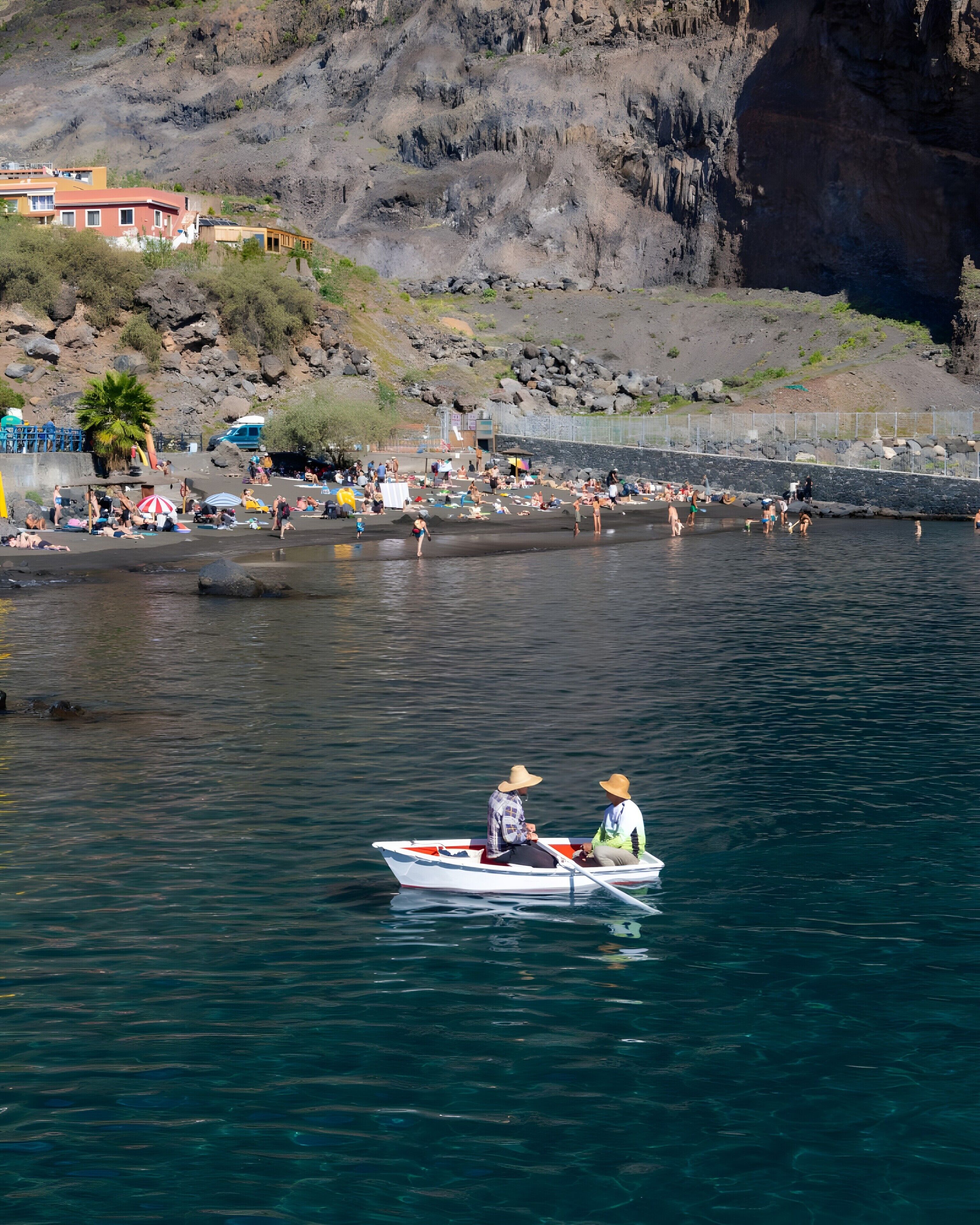 Un paysage côtier avec une falaise rocheuse, une plage où se trouvent des personnes, et un bateau avec deux personnes à bord.