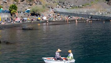 Un paysage côtier avec une falaise rocheuse, une plage où se trouvent des personnes, et un bateau avec deux personnes à bord.