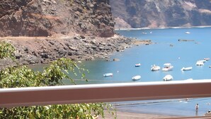 Un balcón con vistas a una costa rocosa, barcos en el agua y una mesa con fruta y una copa.