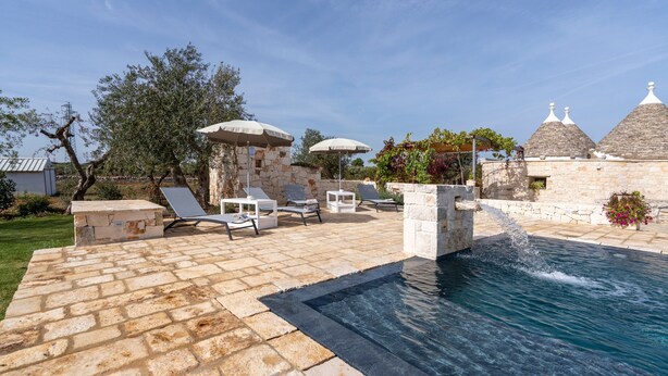 A stone pool with a water feature, lounge chairs, and umbrellas in a courtyard. - Villa 'Trulli Orma' with Private Pool, Wi-Fi and Air Conditioning (Locorotondo)