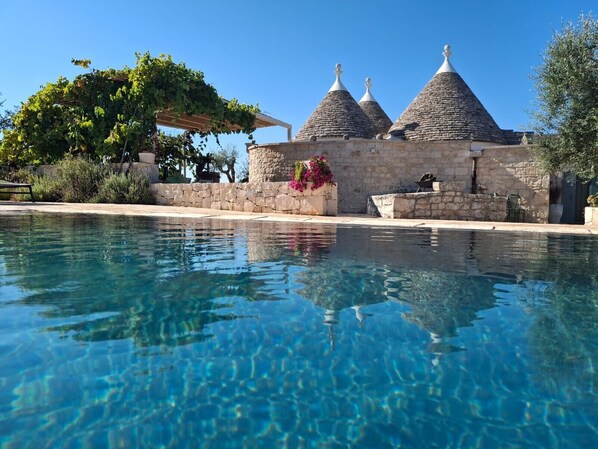 A swimming pool with clear water, a stone building with conical roofs, and a pergola with greenery.