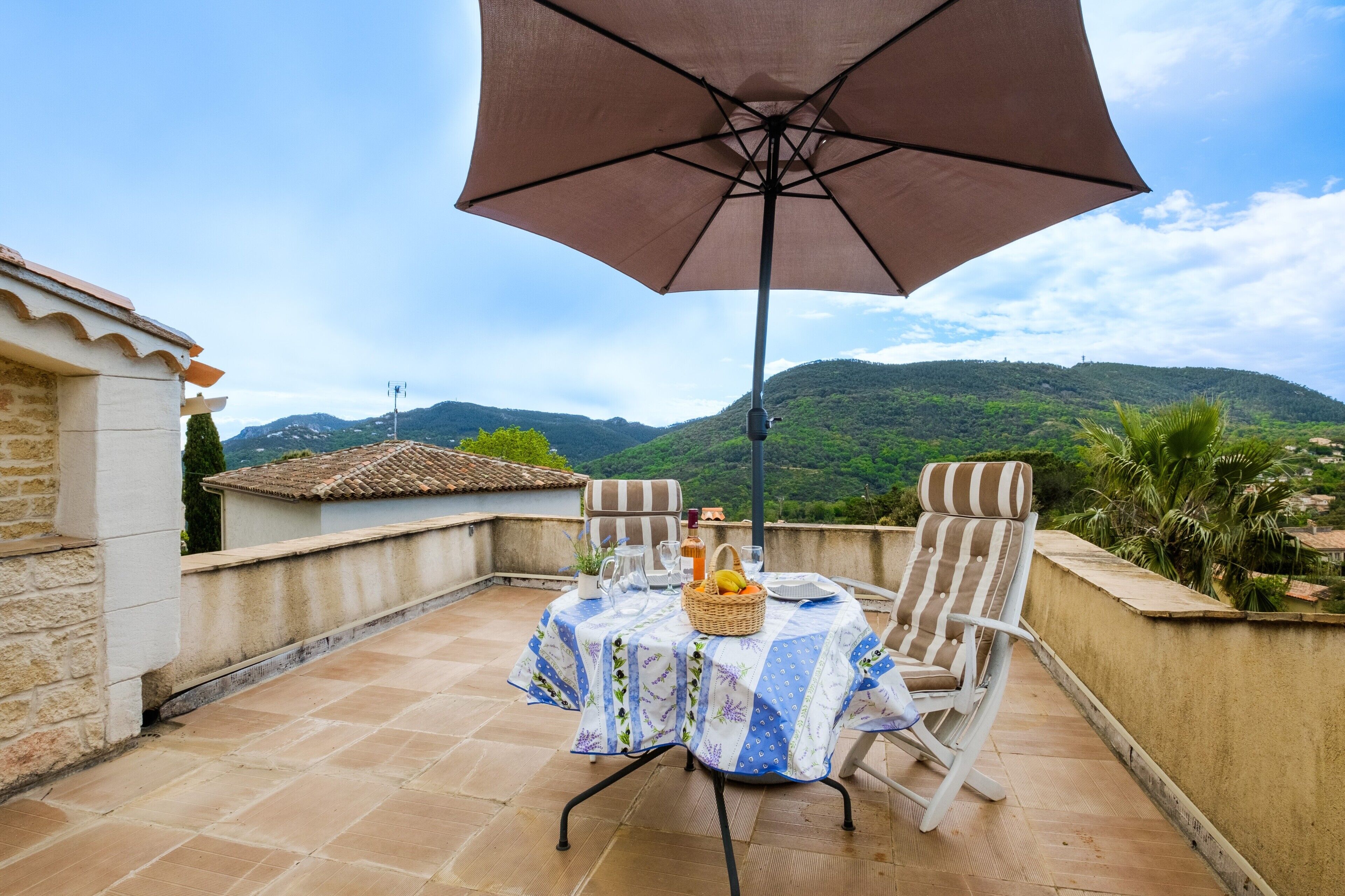 Une terrasse avec une table dressée pour deux personnes, un grand parasol et une vue sur des collines verdoyantes.