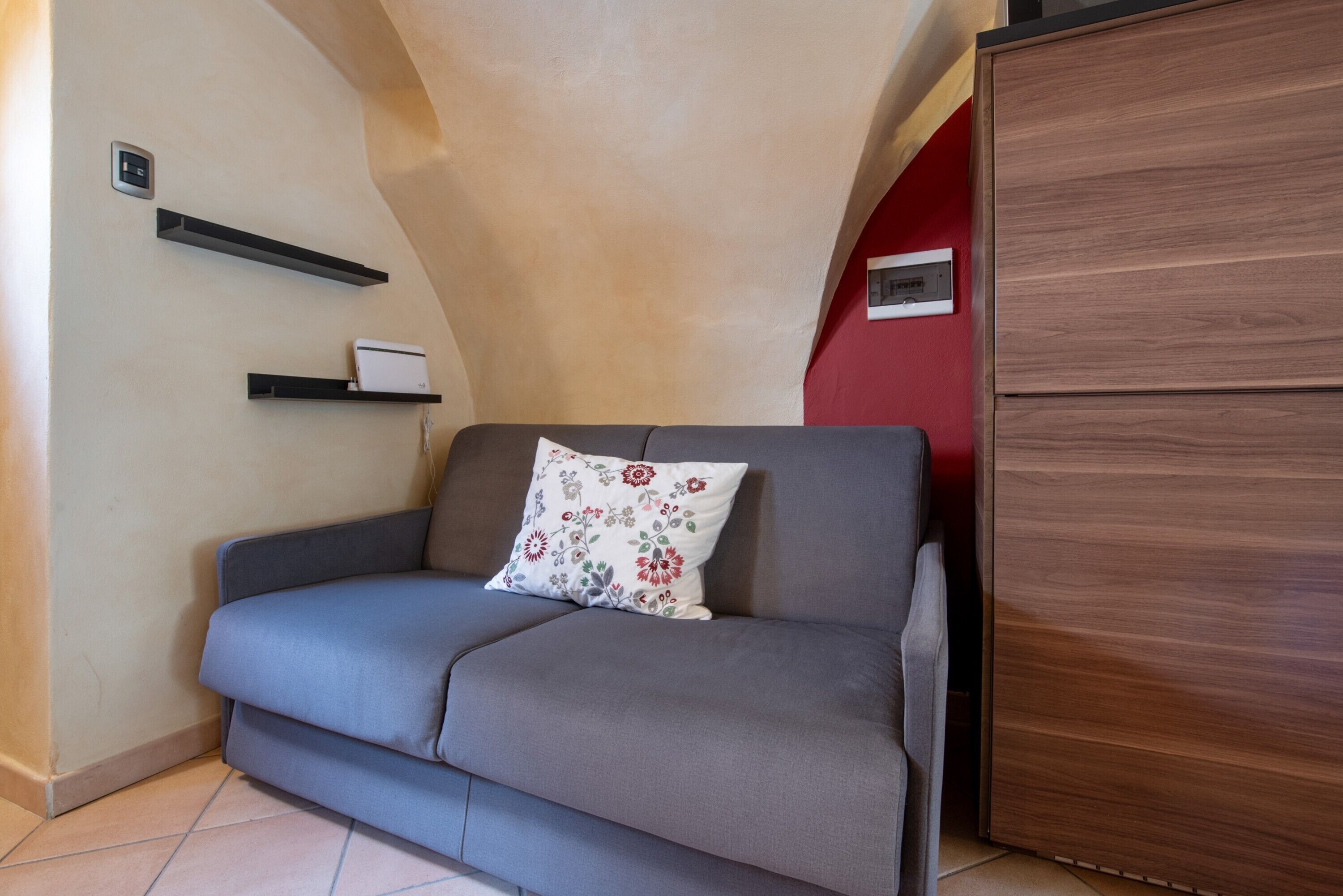 A grey sofa with a floral pillow, a wooden cabinet, and a wall-mounted shelf with a small device.