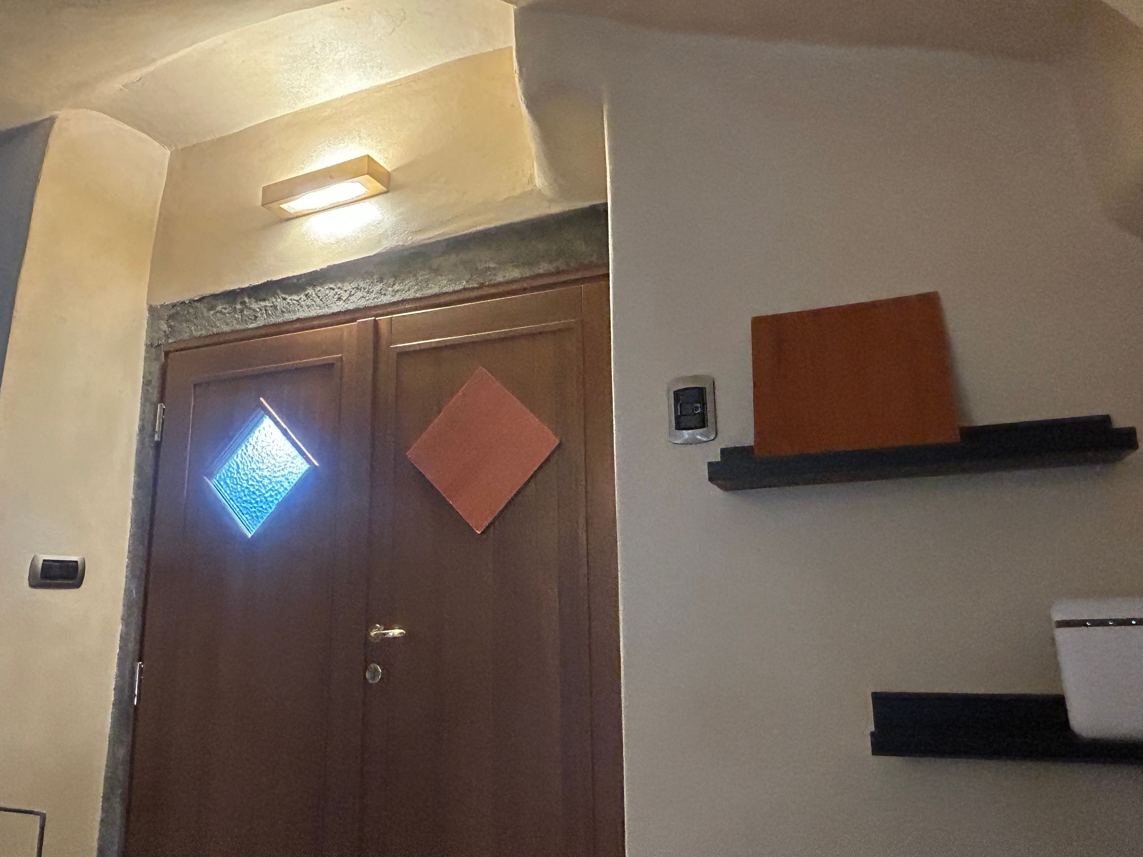 A room with a wooden door, a light fixture above it, and a wall-mounted shelf with a small item on it.