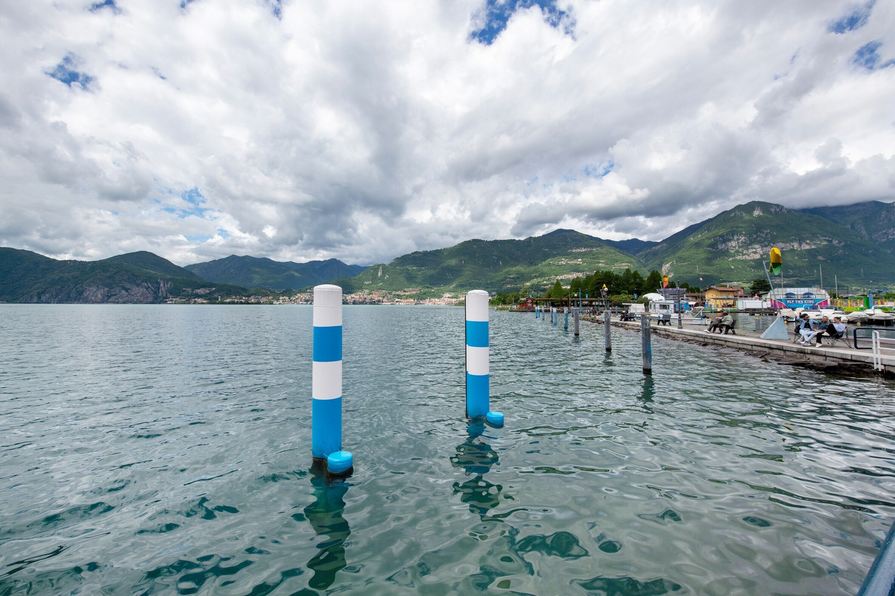 A calm lake with two blue and white poles, a dock with people, and mountains in the background.