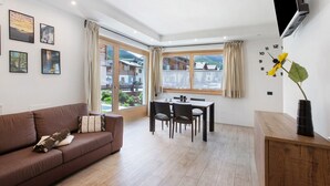 A modern living room with a brown sofa, dining table, and chairs. There is a TV mounted on the wall, a window with curtains, and framed pictures on the wall. - Apartment 'Chalet Snowlake Neve' with Mountain View, Private Garden and Wi-Fi (Livigno)