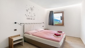 A bedroom with a bed, a nightstand, a wall-mounted heater, a window with curtains, and a view of mountains. - Apartment 'Chalet Snowlake Neve' with Mountain View, Private Garden and Wi-Fi (Livigno)
