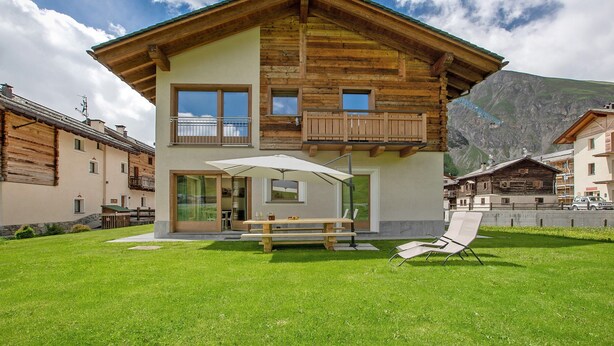 A two-story wooden house with a balcony, a patio with a table and chairs, and a mountain backdrop. - Apartment 'Chalet Snowlake Neve' with Mountain View, Private Garden and Wi-Fi (Livigno)