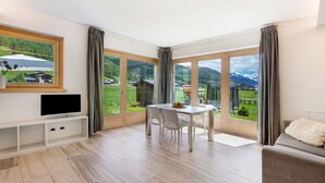 Een moderne kamer met een eettafel, stoelen, een tv op een standaard en grote ramen voorzien van gordijnen.