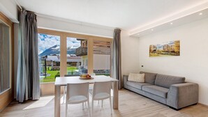 Een moderne woonkamer met een eettafel, twee stoelen, een grijze bank en een groot raam met uitzicht op de bergen.