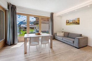 Un salon moderne comprenant une table à manger, deux chaises, un canapé gris et une grande fenêtre donnant sur une vue imprenable sur les montagnes.