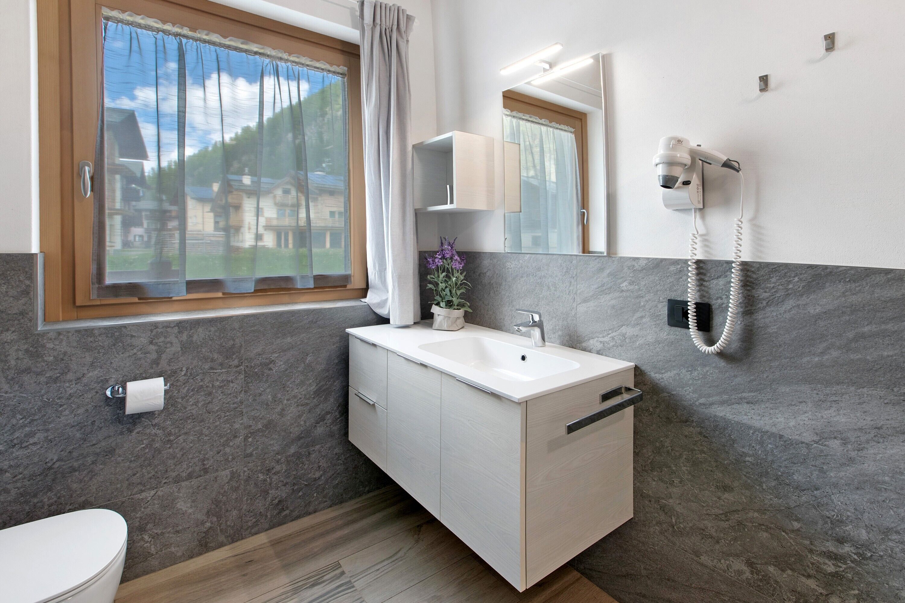 A bathroom with a toilet, a sink, a mirror, a shower, and a window with a view of houses.