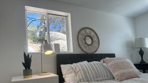 Une chambre à coucher avec un lit, une table de chevet avec une lampe, une fenêtre donnant sur des arbres et un objet décoratif fixé au mur.