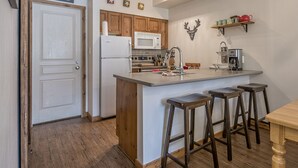 A kitchen with wooden cabinets, a white refrigerator, a microwave, a coffee machine, and a bar stool seating area. - Pet friendly ground floor condo with mudroom directly slope side at Creekside (Silver Star Mountain)