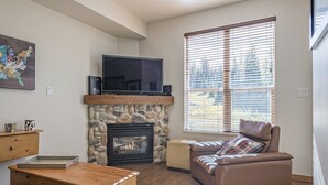 A living room with a stone fireplace, a flat-screen TV, a leather sofa, and a large window with blinds. - Pet friendly ground floor condo with mudroom directly slope side at Creekside (Silver Star Mountain)