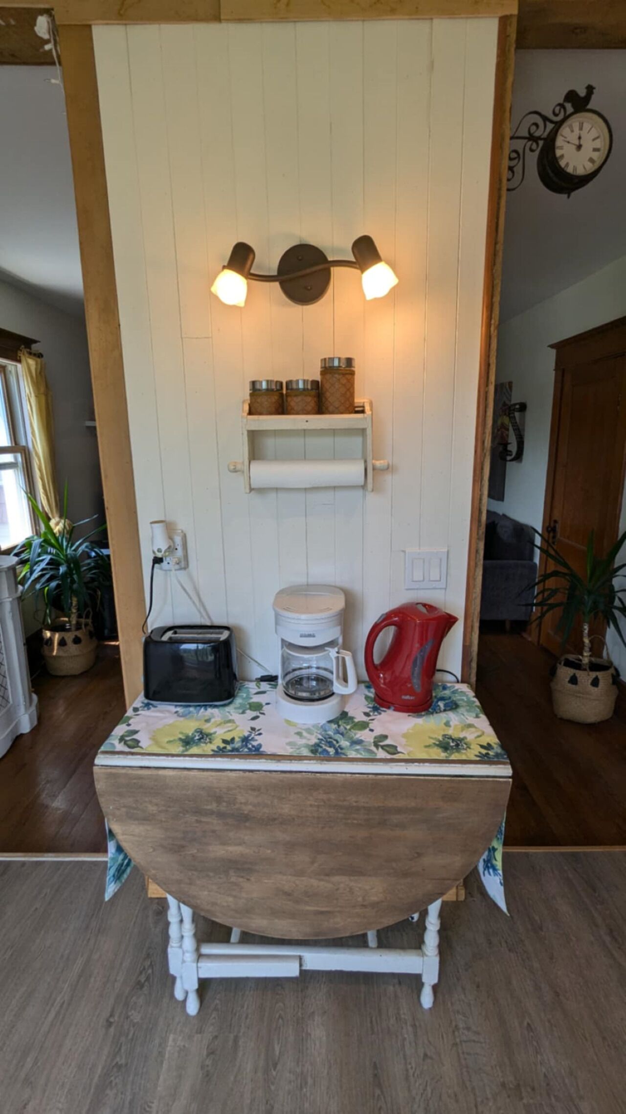 A rustic kitchen with a wooden table, a coffee maker, a kettle, and a shelf with jars.