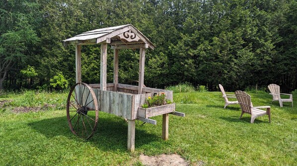 Une charrette en bois rustique, dotée d’un toit, est placée sur une pelouse, à proximité de deux fauteuils Adirondack.