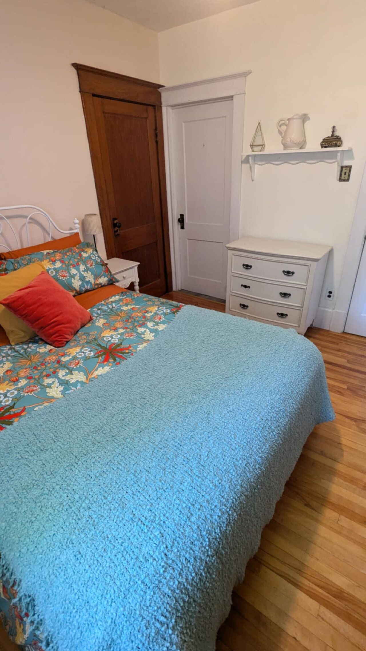 Een slaapkamer met een bed, nachtkastje, dressoir, deur en een plank met decoratieve objecten.
