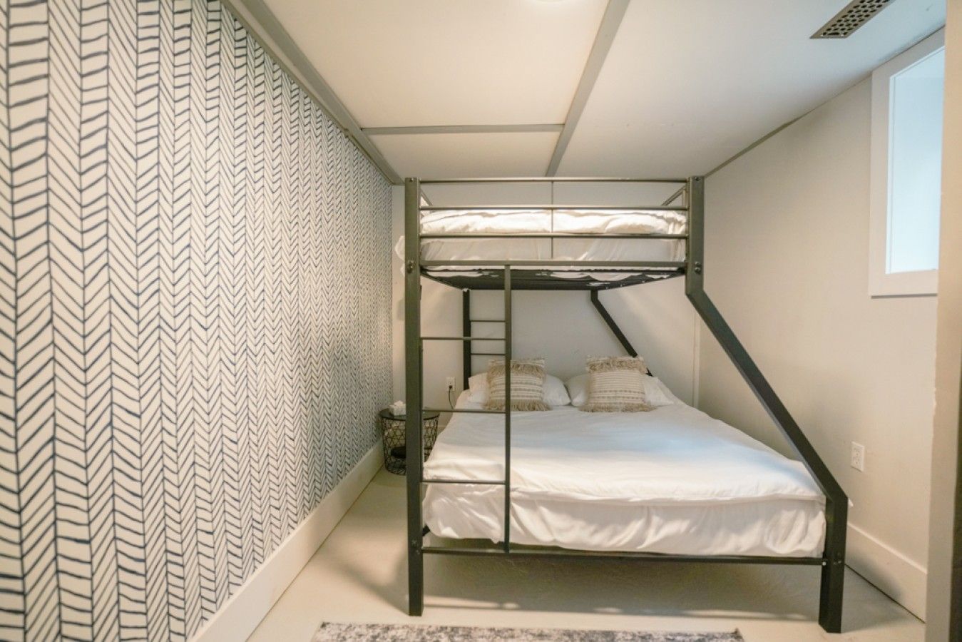 Ein Stockbett in einem Zimmer mit ornamentalem Wandpapier, ein kleiner Nachttisch und ein Fenster.