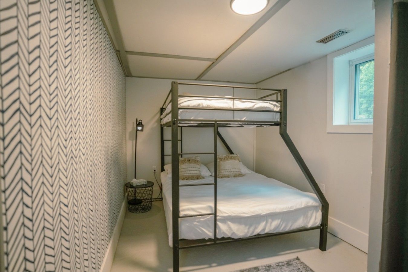 A bunk bed with a ladder, a small table with a lamp, and a window with daylight.